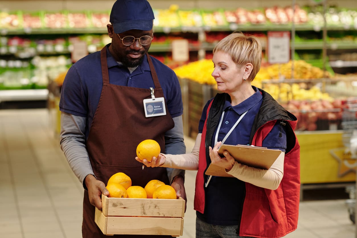 Two supermarket workers stand in the produce section. One holds a crate of oranges while the other, next to a commercial citrus juicer, examines an orange and holds a clipboard. Shelves with fruits and vegetables are visible in the background.