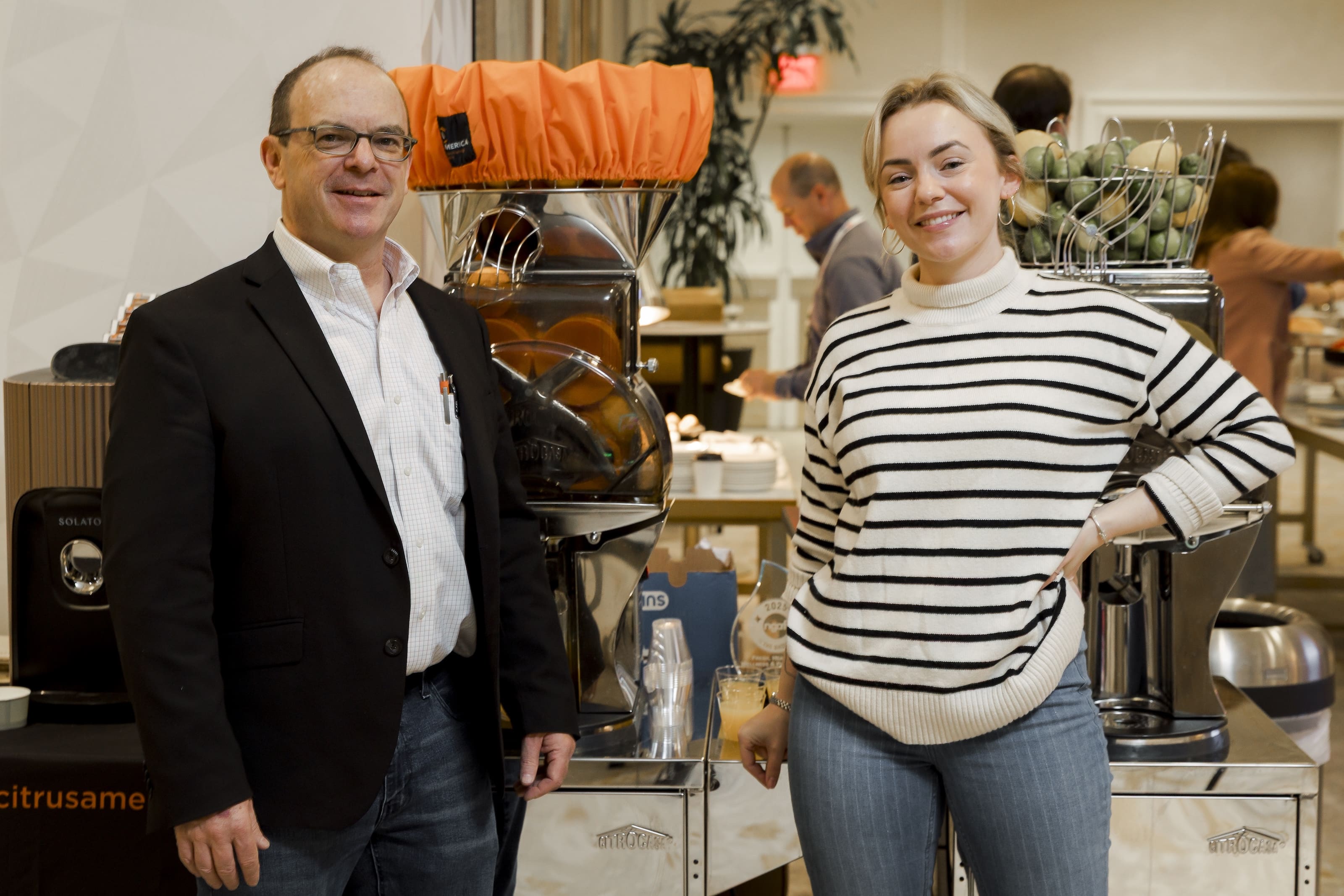 A man in a blazer and a woman in a striped sweater stand smiling next to two juicing machines at an indoor event, showcasing Fresh Juice Innovation for Retailers, with other people and tables visible in the background.
